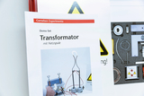 Das Bild zeigt eine Anleitung für ein Demo-Set zum Thema Transformator mit Netzspule. Im Hintergrund sind verschiedene experimentelle Geräte und Sicherheitswarnungen zu sehen, die auf eine experimentelle Nutzung hinweisen.