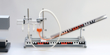 Das Bild zeigt ein Experimentiergerät mit einer schrägen Röhre, die eine Flüssigkeit in eine durchsichtige Kammer leitet. Neben der Röhre befinden sich verschiedene Anschlüsse und ein elektrisches Gerät, das vermutlich zur Steuerung des Experiments dient.
