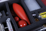 Das Bild zeigt einen Koffer mit Experimentiermaterialien, die zur Durchführung von wissenschaftlichen Tests geeignet sind. Darin befinden sich verschiedene rote Objekte, darunter eine längliche Form und mehrere Kugeln, sowie einige weitere Utensilien.
