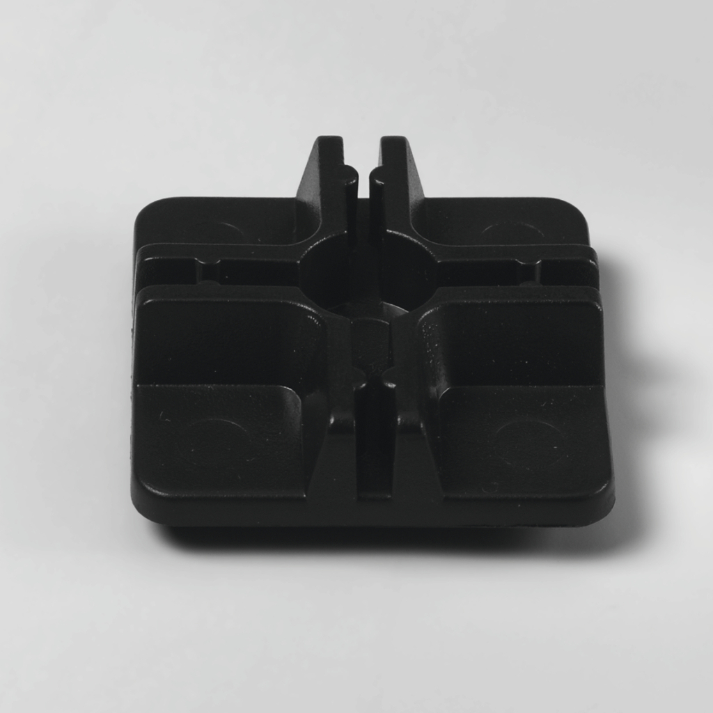 Das Bild zeigt ein quadratisches, schwarzes Kunststoffteil mit abgerundeten Ecken und vertieften Bereichen. Es scheint sich um eine Halterung oder Basis zu handeln, die möglicherweise für experimentelle Setups im Bildungsbereich verwendet wird.