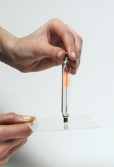 Das Bild zeigt eine Hand, die eine gläserne Röhre mit einer leuchtenden Spitze über einer transparenten Platte hält. Diese Apparatur könnte für Experimente zur Demonstration von elektrischen Entladungen oder Plasma verwendet werden.