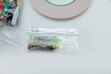 Das Bild zeigt eine transparente Tüte mit verschiedenen farbigen LEDs (rot, gelb, grün) und einem Pack mit elektrischen Leitungen in verschiedenen Farben. Darüber hinaus ist eine Rolle doppelseitiges Klebeband zu sehen, die für Experimentierprojekte verwendet werden kann.