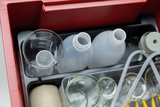 Das Bild zeigt eine Sammlung von Experimentiergeräten, darunter mehrere Messbecher, Flaschen und Schläuche, die in einem roten Kasten angeordnet sind. Diese Materialien sind typisch für den Chemieunterricht und die Durchführung wissenschaftlicher Experimente.