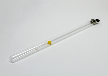 Das Bild zeigt ein transparentes Glasröhrchen mit einer gelben Kugel im Inneren. Das Röhrchen hat an einem Ende einen Anschluss, wahrscheinlich für Experimentierzwecke im Bildungsbereich.