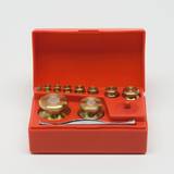 Das Bild zeigt ein Set von goldfarbenen Gewichten in einem roten Kunststoffkoffer. Die Gewichte variieren in Größe und sind für experimentelle Zwecke im Bildungswesen vorgesehen.