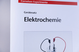 Das Bild zeigt ein Produkt aus einem Online-Shop für Experimentierbedarf, speziell für das Thema Elektrochemie. Es illustriert einen Gerätesatz mit einer Abbildung eines Elektrolyseversuchs, inklusive einer transparenten Zelle und Kabeln in Schwarz und Rot.