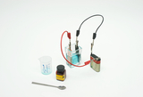 Das Bild zeigt ein Experimentierset mit einem Elektrolysegerät, bestehend aus einer transparenten Kammer mit blauer Flüssigkeit, zwei Kabeln, einer Batterie sowie einem Messbecher und einem Reagenzglas. Daneben befindet sich ein Behälter mit chemischem Reagenz und ein Löffel.