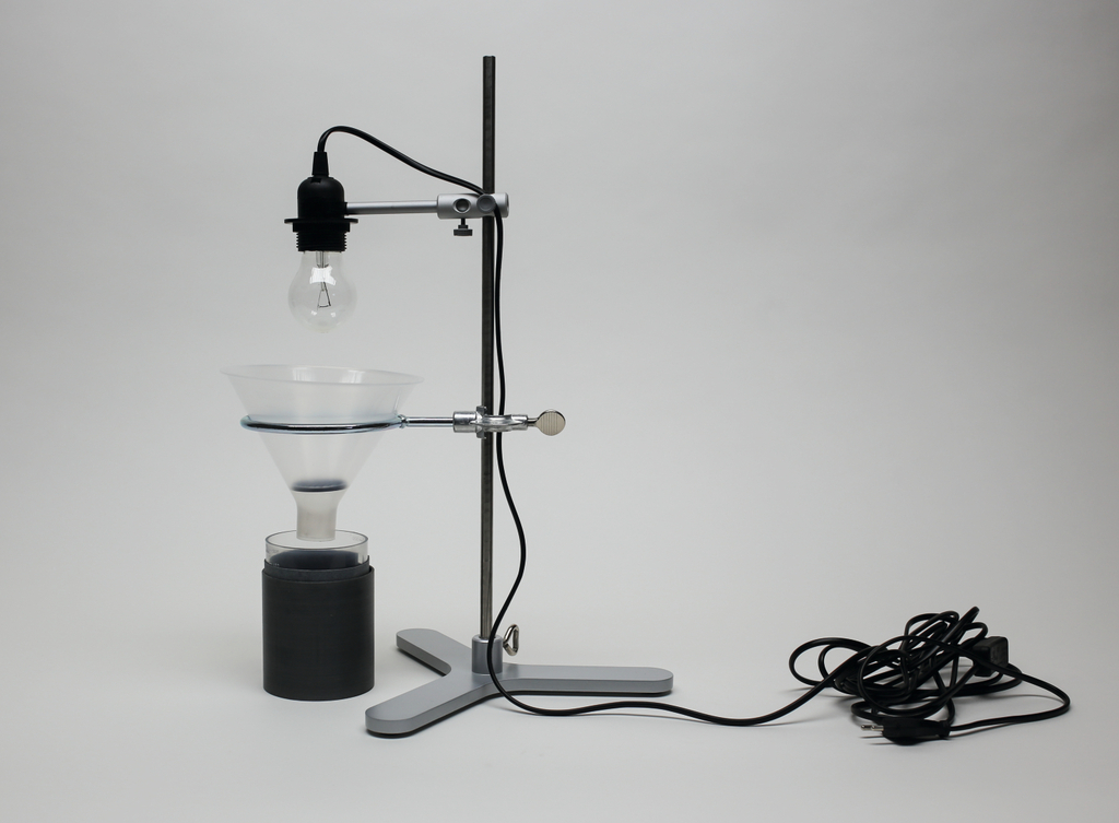Das Bild zeigt ein Experimentiergerät mit einer Lampenfassung, die über einem Trichter montiert ist. Darunter befindet sich ein schwarzer Behälter, der wahrscheinlich für chemische Experimente oder als Teil eines physikalischen Versuchs verwendet wird.