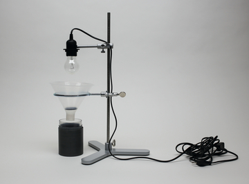 Das Bild zeigt ein Experimentiergerät mit einer Lampenfassung, die über einem Trichter montiert ist. Darunter befindet sich ein schwarzer Behälter, der wahrscheinlich für chemische Experimente oder als Teil eines physikalischen Versuchs verwendet wird.