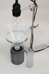 Das Bild zeigt ein Experimentiergerät, das aus einer Halterung mit einer Lichtquelle und einem transparenten Trichter besteht. Unter dem Trichter befindet sich ein Zylinder, der vermutlich zur Aufnahme von Flüssigkeiten oder Gasen dient.