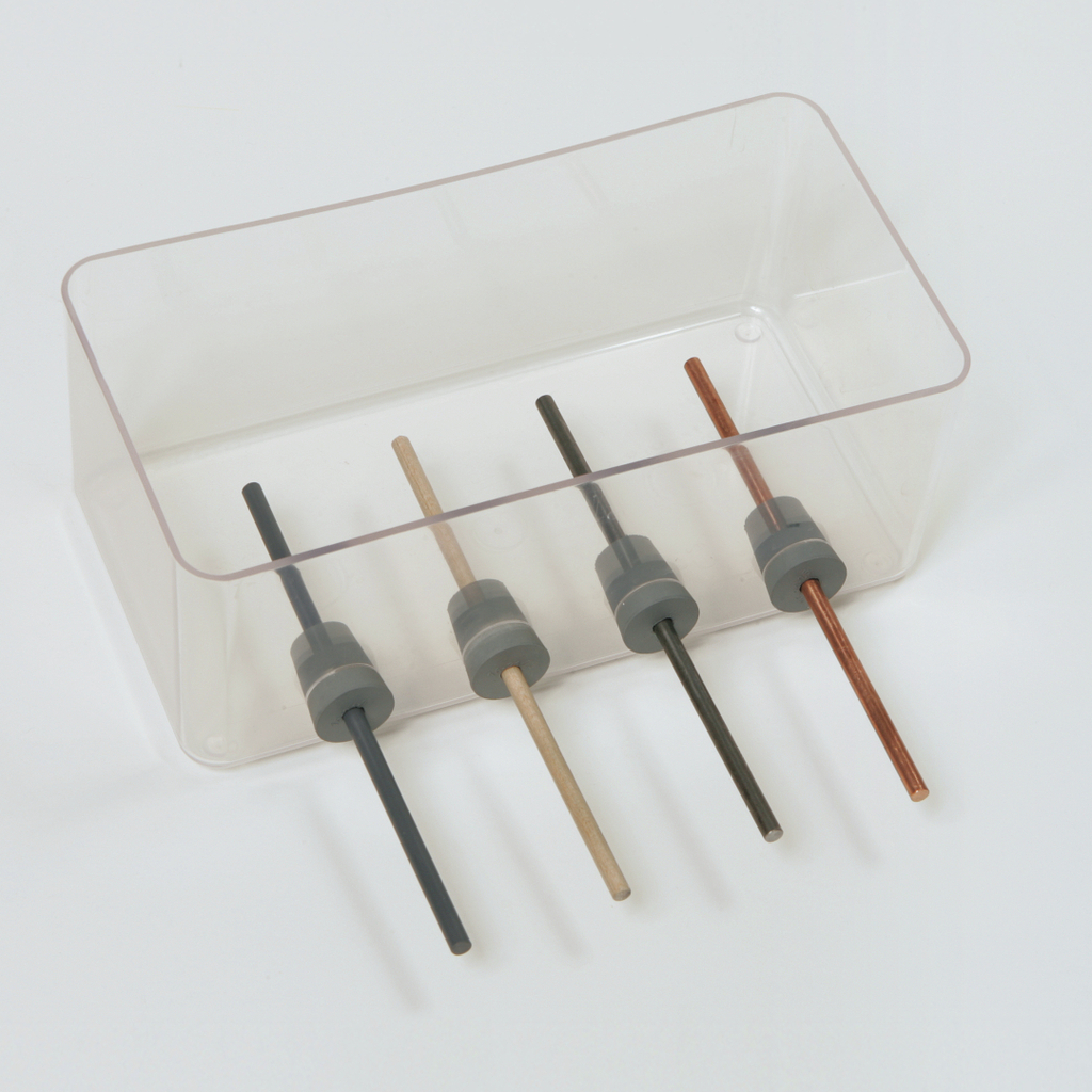 Das Bild zeigt eine transparente Box mit vier unterschiedlichen Stäben, die in grauen Haltern fixiert sind. Die Stäbe bestehen aus verschiedenen Materialien und Farben, möglicherweise für experimentelle Zwecke im Bildungsbereich.