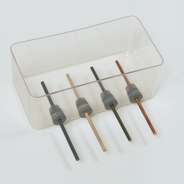 Das Bild zeigt eine transparente Box mit vier unterschiedlichen Stäben, die in grauen Haltern fixiert sind. Die Stäbe bestehen aus verschiedenen Materialien und Farben, möglicherweise für experimentelle Zwecke im Bildungsbereich.
