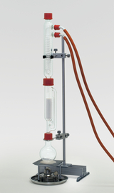 Das Bild zeigt ein chemisches Experimentiergerät, bestehend aus mehreren Glasbehältern und Schläuchen, die auf einem stabilen Standfuß montiert sind. Unten befindet sich eine Heizquelle, während das Setup zur Durchführung von chemischen Reaktionen oder Destillationen dienen kann.