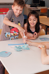Auf dem Bild experimentieren Kinder in einem Klassenraum mit einem wissenschaftlichen Experiment. Ein Junge verwendet eine Spritze, während ein Mädchen gespannt zuschaut, und auf dem Tisch liegen Materialien in einem Plastikbeutel.