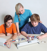 Das Bild zeigt drei Schüler, die an einem Experimentierapparat arbeiten, der mit Elektromotoren und Propellern ausgestattet ist. Sie sind konzentriert bei der Durchführung des Versuchs, der wahrscheinlich physikalische Konzepte wie Bewegung und Antrieb vermittelt.