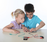 Das Bild zeigt zwei Kinder, die konzentriert an einem Experiment mit Batterien und Drähten arbeiten. Sie nutzen Materialien, um einen Schaltkreis zu erstellen, was eine spielerische Einführung in die Grundlagen der Elektrizität darstellt.