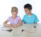 Das Bild zeigt zwei Kinder, die an einem Experiment mit elektrischen Schaltkreisen arbeiten. Das Mädchen verbindet Kabel mit einer Batterie und einer Lampe, während der Junge interessiert zuschaut.