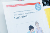 Das Bild zeigt eine Broschüre von Cornelsen Experimente, die sich mit naturwissenschaftlichen Experimenten im Kindergarten befasst. Der Titel thematisiert das Experimentieren mit Elektrizität durch die Kinder Anja und Leon.