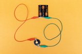 Das Bild zeigt eine Anordnung von zwei Batterien, die durch farbige Kabel mit einem Kontakt verbunden sind. Diese Konfiguration dient vermutlich zur Durchführung elektrischer Experimente im Bildungsbereich.
