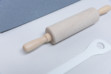 Das Bild zeigt einen Holzrolling Pin, der zentral auf weißem Hintergrund platziert ist, zusammen mit einem weißen Werkzeug und einem blauen Materialstück im Hintergrund. Diese Gegenstände eignen sich gut für experimentelle Aktivitäten im Bildungsbereich.