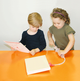 Auf dem Bild sind zwei Kinder an einem orangefarbenen Tisch zu sehen. Das eine Kind hält ein Blatt Papier, während das andere mit einem Experimentiergerät arbeitet, das an einem offenen Buch befestigt ist.