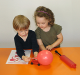 Das Bild zeigt zwei Kinder, die gemeinsam an einem Experiment mit einem roten Luftballon und einem kleinen Wagen arbeiten. Auf dem Tisch liegt ein Arbeitsblatt, und neben den Kindern befindet sich eine Luftpumpe.
