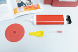 Das Bild zeigt verschiedene Experimentiermaterialien, darunter eine rote Pumpe, einen gelben Ballon und eine rote Scheibe. Zusätzlich ist ein Buch mit Anleitungen zu Experimenten abgebildet, das die Verwendung der Materialien erklärt.