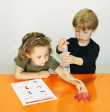 Auf dem Bild sind zwei Kinder zu sehen, die an einem Experiment arbeiten. Das Mädchen folgt einer Anleitung auf einem Papier, während der Junge mit einer Spritze eine Flüssigkeit in ein Gerät leitet.
