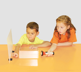 Das Bild zeigt zwei Kinder, die auf einem orangefarbenen Tisch experimentieren. Das Mädchen hält eine Taschenlampe, während der Junge auf eine durchsichtige Box und eine reflektierende Fläche zeigt, was auf ein spannendes Lernexperiment hindeutet.