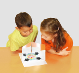 Das Bild zeigt zwei Kinder, einen Jungen in einem gelben T-Shirt und ein Mädchen in einem orangefarbenen T-Shirt, die neugierig auf ein Experimentiervorhaben schauen. Vor ihnen befindet sich eine aufgestellte Fläche mit Spiegeln und verschiedenen Objekten, die zum Experimentieren einladen.