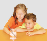 Das Bild zeigt ein Mädchen und einen Jungen, die gebannt auf ein Glas mit Wasser und Eiswürfeln schauen. Sie experimentieren offensichtlich und beobachten, wie ein grauer Strohhalm im Glas platziert ist.