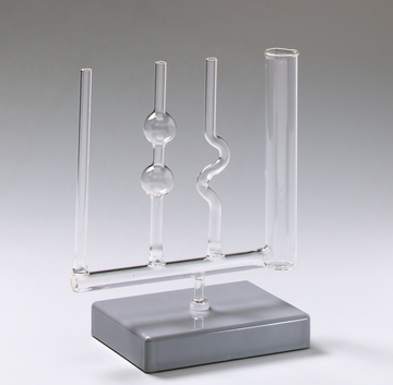 Das Bild zeigt ein Labormaterial mit mehreren Glasröhren, die auf einem grauen Standfuß befestigt sind. Die Röhren haben unterschiedliche Formen, darunter eine schlangenförmige, und scheinen für chemische Experimente oder Demonstrationen konzipiert zu sein.