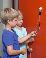 Auf dem Bild sind zwei Kinder zu sehen, die an einem Experiment mit einer Aufhängung und einer Schnur arbeiten. Die orangefarbene Wand im Hintergrund schafft einen lebhaften Kontrast zu den Aktivitäten der Kinder, die neugierig und konzentriert wirken.