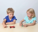 Auf dem Bild sind zwei Kinder zu sehen, die an einem Tisch sitzen und mit Spielzeugfahrzeugen experimentieren. Das Mädchen in der blauen T-Shirt lächelt, während der Junge konzentriert auf sein rotes Fahrzeug schaut.