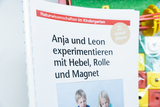Das Bild zeigt eine Seite eines Buches mit dem Titel „Naturwissenschaften im Kindergarten“, auf dem Kinder namens Anja und Leon abgebildet sind, die mit Hebel, Rolle und Magnet experimentieren. Im Hintergrund sind verschiedene Experimentiermaterialien sichtbar, die für den Bildungsbereich gedacht sind.