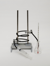 Das Bild zeigt eine experimentelle Einrichtung, die aus einem Stativ mit zwei metallischen Stangen, einem Spiralrohr, einer Spritze und einem Erlenmeyerkolben mit einem Thermometer besteht. Diese Ausrüstung wird typischerweise für chemische Experimente im Bildungswesen verwendet.