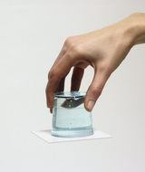 Auf dem Bild ist eine Hand zu sehen, die ein Glas mit Wasser anhebt, welches auf einem weißen Blatt Papier steht. Die Szenerie vermittelt einen experimentellen Charakter, möglicherweise für Demonstrationen in pädagogischen Kontexten.