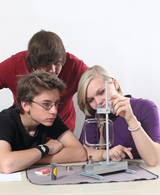 Das Bild zeigt drei Jugendliche, die konzentriert an einem Experimentieraufbau arbeiten. Vor ihnen steht ein Apparatur mit einer transparenten Kammer, während sie verschiedene Materialien und Werkzeuge auf einem Tisch untersuchen.