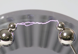 Das Bild zeigt zwei metallische Kugeln, die durch einen elektrischen Funken verbunden sind. Dieser Funke illustriert ein physikalisches Experiment, das häufig in der Schulausbildung verwendet wird, um Konzepten der Elektrizität und des Stroms zu veranschaulichen.