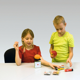 Auf dem Bild sind zwei Kinder an einem Tisch zu sehen, die mit verschiedenen Objekten experimentieren. Ein Kind hält einen Apfel und das andere eine Orange auf einer Waage, während sie weitere Materialien wie Stifte und Kartenspiele untersuchen.