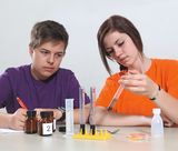 Das Bild zeigt zwei Jugendliche, die an einem Experiment teilnehmen. Die junge Frau in einem orangenen T-Shirt hält ein Reagenzglas, während der Junge in lila T-Shirt aufmerksam zuschaut; auf dem Tisch befinden sich verschiedene Behälter und Messgeräte.