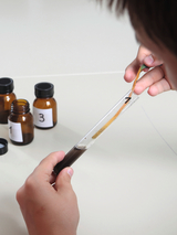 Das Bild zeigt eine Hand, die eine Reagenzglasprobe hält, während ein Streifen in die Flüssigkeit eingetaucht wird. Im Hintergrund sind mehrere braune Fläschchen mit Nummern zu sehen, die auf verschiedene Chemikalien oder Lösungen hindeuten.