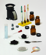 Das Bild zeigt eine Sammlung von Experimentiermaterialien, darunter Messgeräte, Reagenzgläser mit verschiedenen Flüssigkeiten, und Gläser mit Pulver. Außerdem sind Pipetten und eine Farbskala zur pH-Wertbestimmung zu sehen, die für chemische Experimente verwendet werden können.