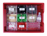 Das Bild zeigt eine rote Aufbewahrungsbox, die mit verschiedenen Materialien für Experimente gefüllt ist, darunter Pappe, Kunststoffe, Holzstäbchen und Baumwollstoffe. Zusätzlich sind ein Fläschchen mit Tafelessig und mehrere transparente Behälter enthalten.