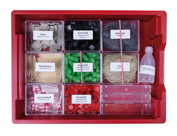 Das Bild zeigt eine rote Aufbewahrungsbox, die mit verschiedenen Materialien für Experimente gefüllt ist, darunter Pappe, Kunststoffe, Holzstäbchen und Baumwollstoffe. Zusätzlich sind ein Fläschchen mit Tafelessig und mehrere transparente Behälter enthalten.