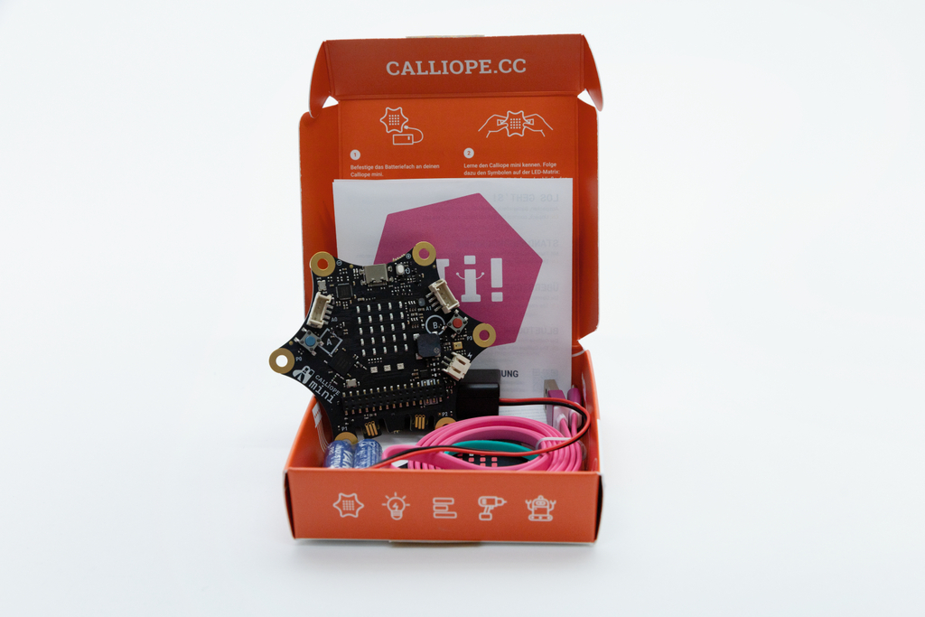 Das Bild zeigt ein Calliope Mini-Board in seiner orangefarbenen Verpackung, umgeben von verschiedenen Kabeln und einer Anleitung. Das Board dient als leicht zugängliches Werkzeug für Schüler, um Programmierung und Elektronik spielerisch zu lernen.