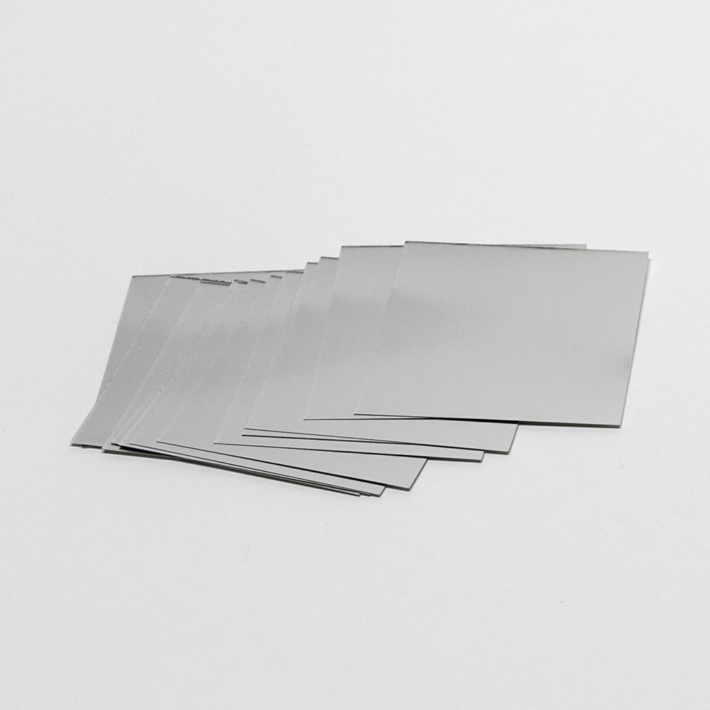 Das Bild zeigt eine Reihe von quadratischen, metallischen Platten, die auf einem weißen Untergrund liegen. Sie scheinen Teil eines Experimentierkits zu sein, das im Rahmen von Bildungszwecken verwendet wird.