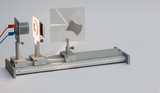 Das Bild zeigt ein physikalisches Experimentiergerät, bestehend aus einer Lichtquelle und einem transparenten Element, das Lichtstrahlen projiziert. Es ist auf einem schienenartigen Gestell montiert, das eine präzise Positionierung ermöglicht.