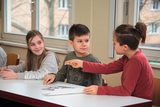 Das Bild zeigt drei Kinder, die an einem Tisch sitzen und miteinander kommunizieren. Die Kinder scheinen engagiert in ein Gespräch vertieft zu sein, während sie auf ein Blatt Papier vor ihnen blicken.