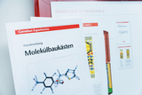 Das Bild zeigt Materialien aus einem Experimentierbedarf für das Bildungswesen, darunter eine Handreichung zu Molekülbaukästen mit einer grafischen Darstellung eines Moleküls. Im Hintergrund ist eine Reihe weiterer Informationsblätter zu sehen.